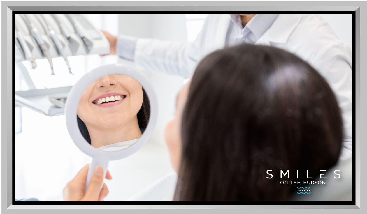Woman smiling while looking in a dental mirror, showcasing a bright smile, with a dentist in the background, emphasizing cosmetic and restorative dentistry at Smiles on the Hudson.