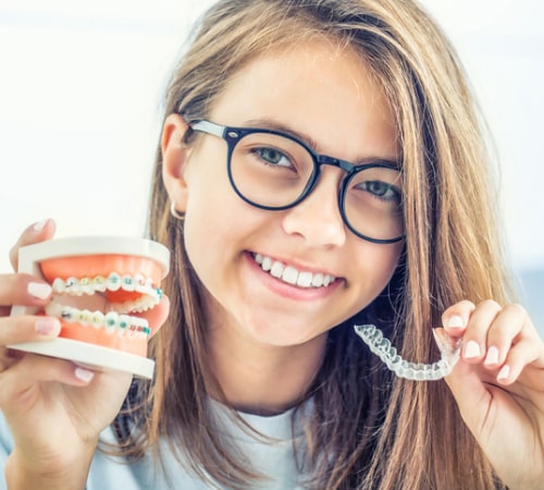 A young woman holds in one hand a teeth model with braces and, in the other, a clear aligner