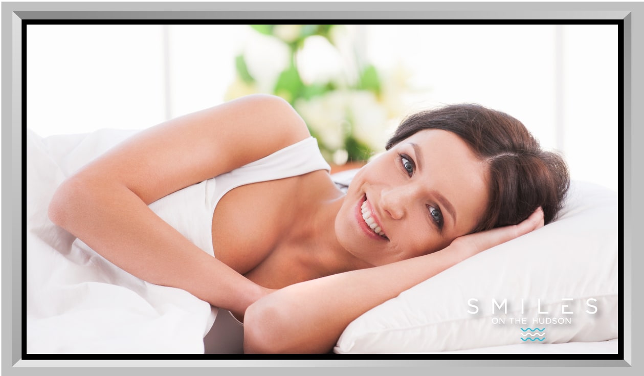 Smiling woman lying on a pillow, representing confidence and comfort in achieving a perfect smile through porcelain veneers, with "Smiles on the Hudson" logo in the corner.